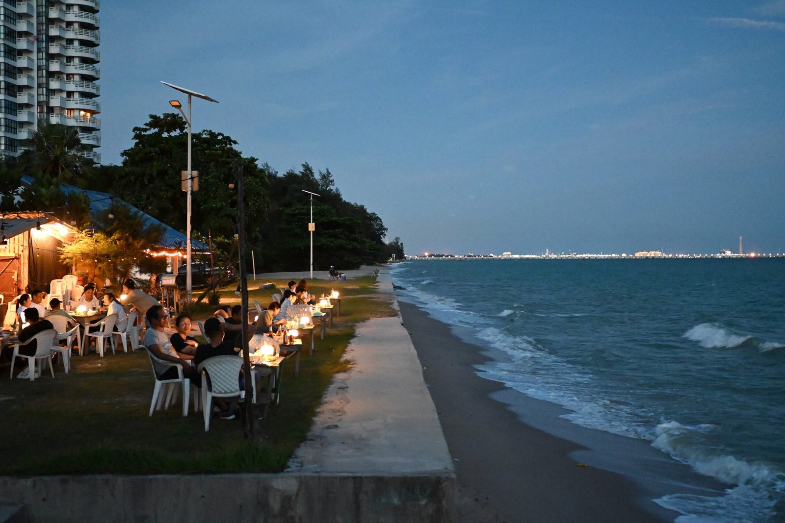 The photo shows a beautiful seaside dining scene at dusk or night. There's an outdoor restaurant or café area with table
