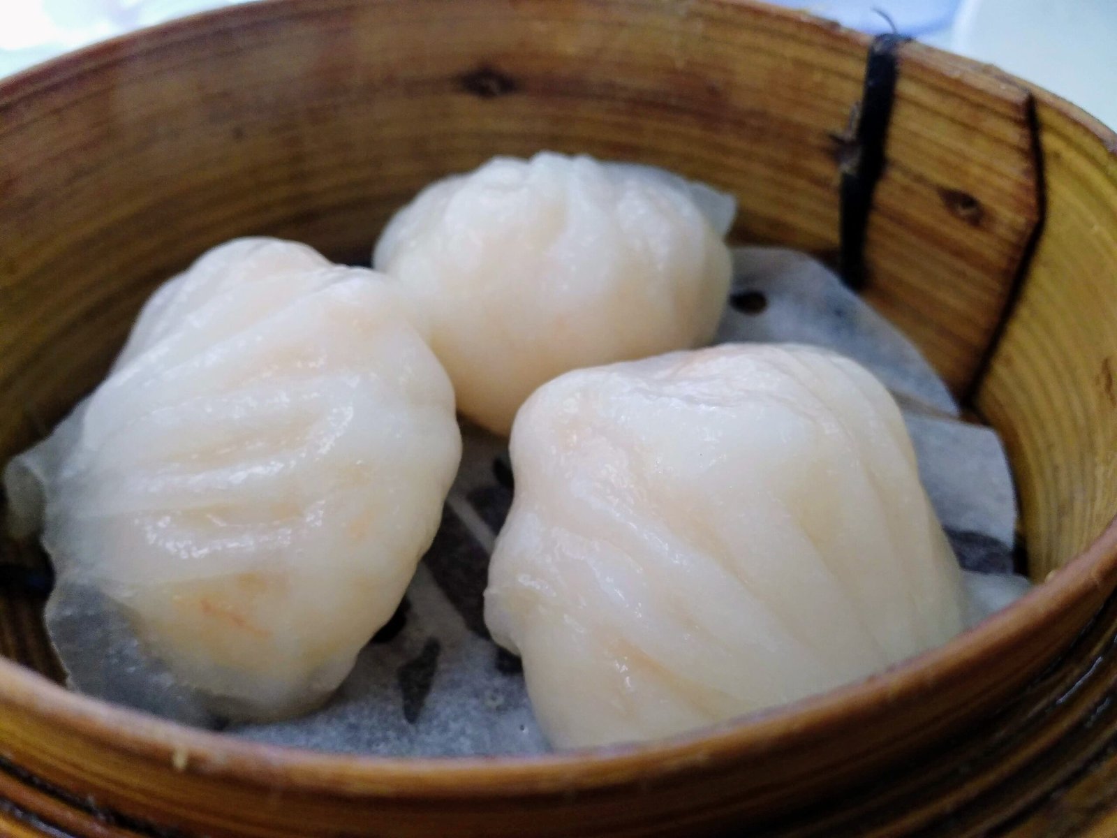 Three pale, peach-colored, wrinkled dim sum in a brown steamer basket resting on white parchment paper lining.